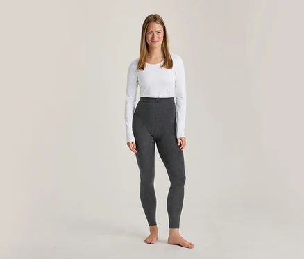 Eine lächelnde Frau steht barfuß in einem weißen Langarmshirt und einer dunkelgrauen, hochtaillierten Leggings.