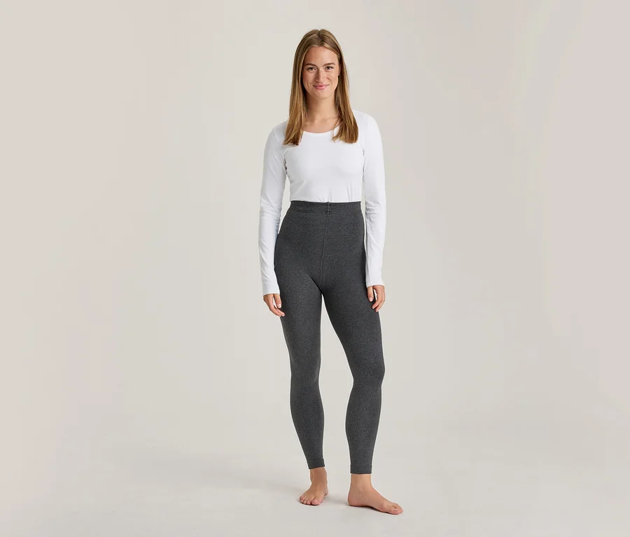 Eine lächelnde Frau steht barfuß in einem weißen Langarmshirt und einer dunkelgrauen, hochtaillierten Leggings.