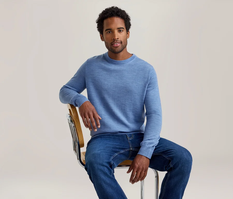 Ein Mann mit dunklem, lockigem Haar trägt einen hellblauen Langarmpullover und blaue Jeans, sitzt auf einem Stuhl und blickt nach vorne.