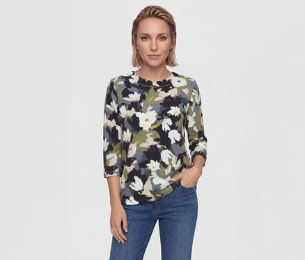 Frau mit kurzen blonden Haaren trägt ein Oberteil mit Blumenmuster in Schwarz, Weiß, Grün, Beige, Grau und blaue Jeans. Eine Hand ist in der Tasche.
