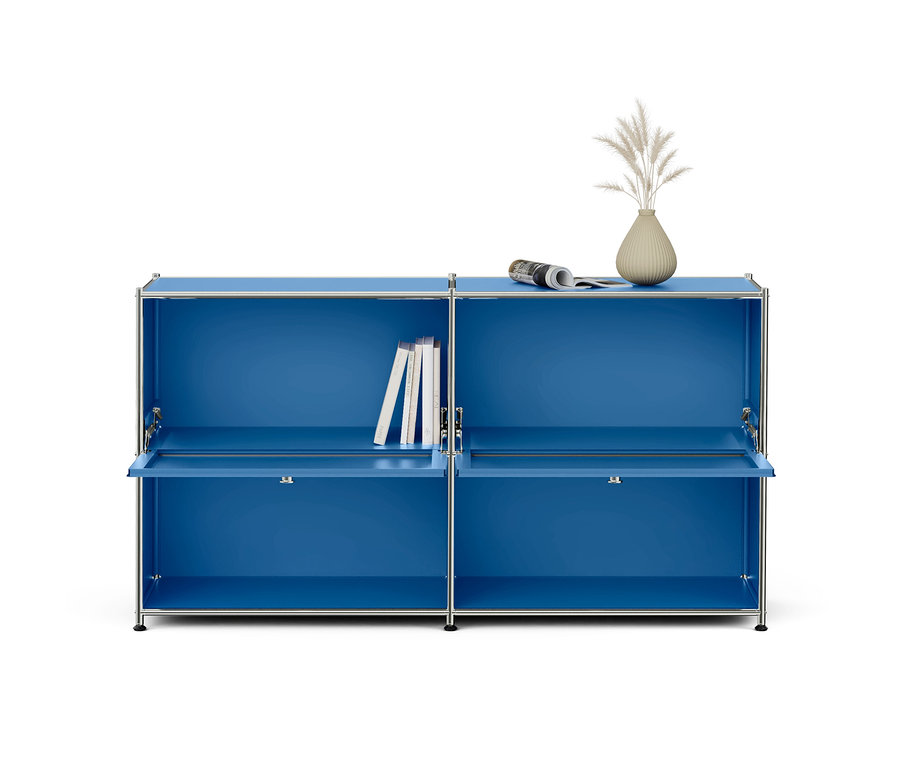 Blauer modularer Schrank mit Chromrahmen, bestehend aus zwei Elementen mit je zwei Fächern. Die unteren Klapptüren sind leicht geöffnet. Im oberen linken Fach stehen Bücher, auf der rechten Oberseite liegen eine Zeitung und eine Vase mit getrockneten Gräsern.