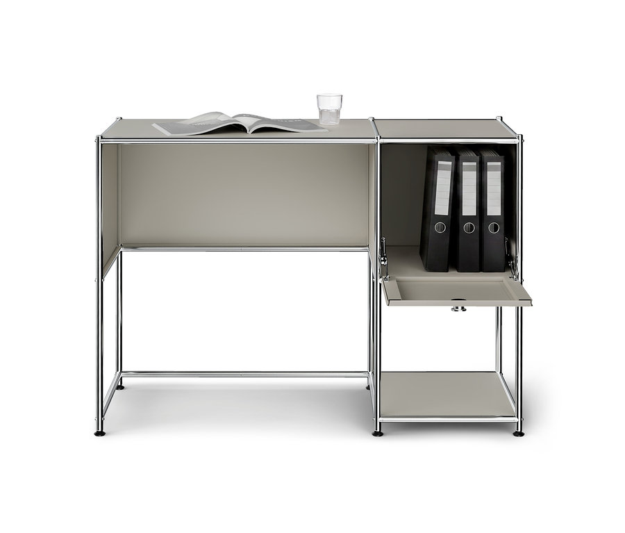 Heller, modularer Schreibtisch in Grau mit Chrombeinen. Auf dem Schreibtisch liegen ein offenes Buch und ein Glas Wasser. Die angebaute Aufbewahrungseinheit rechts enthält drei schwarze Aktenordner, und eine untere Klapptür ist geöffnet, um ein Regal freizulegen.