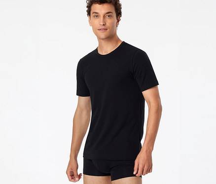 Mann mit lockigem braunem Haar trägt ein schwarzes SCHIESSER T-Shirt und schwarze Boxershorts vor weißem Hintergrund.