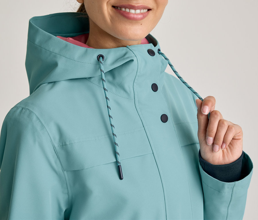 Lächelnde Person in einer türkisfarbenen Kapuzenjacke mit dunklen Knöpfen und pinkem Futter. Ihre Hand zieht an einer Kordel der Kapuze.