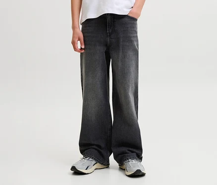 Eine Person trägt dunkelgraue, weit geschnittene Jeans, ein weißes T-Shirt und hellgraue Turnschuhe, eine Hand in der Hosentasche.