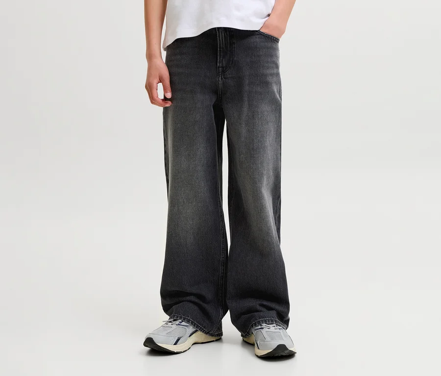 Eine Person trägt dunkelgraue, weit geschnittene Jeans, ein weißes T-Shirt und hellgraue Turnschuhe, eine Hand in der Hosentasche.