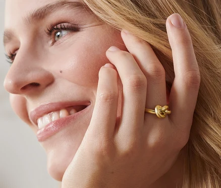 Profil einer lächelnden Blondine mit einem vergoldeten Ring »Knot« am Ringfinger.