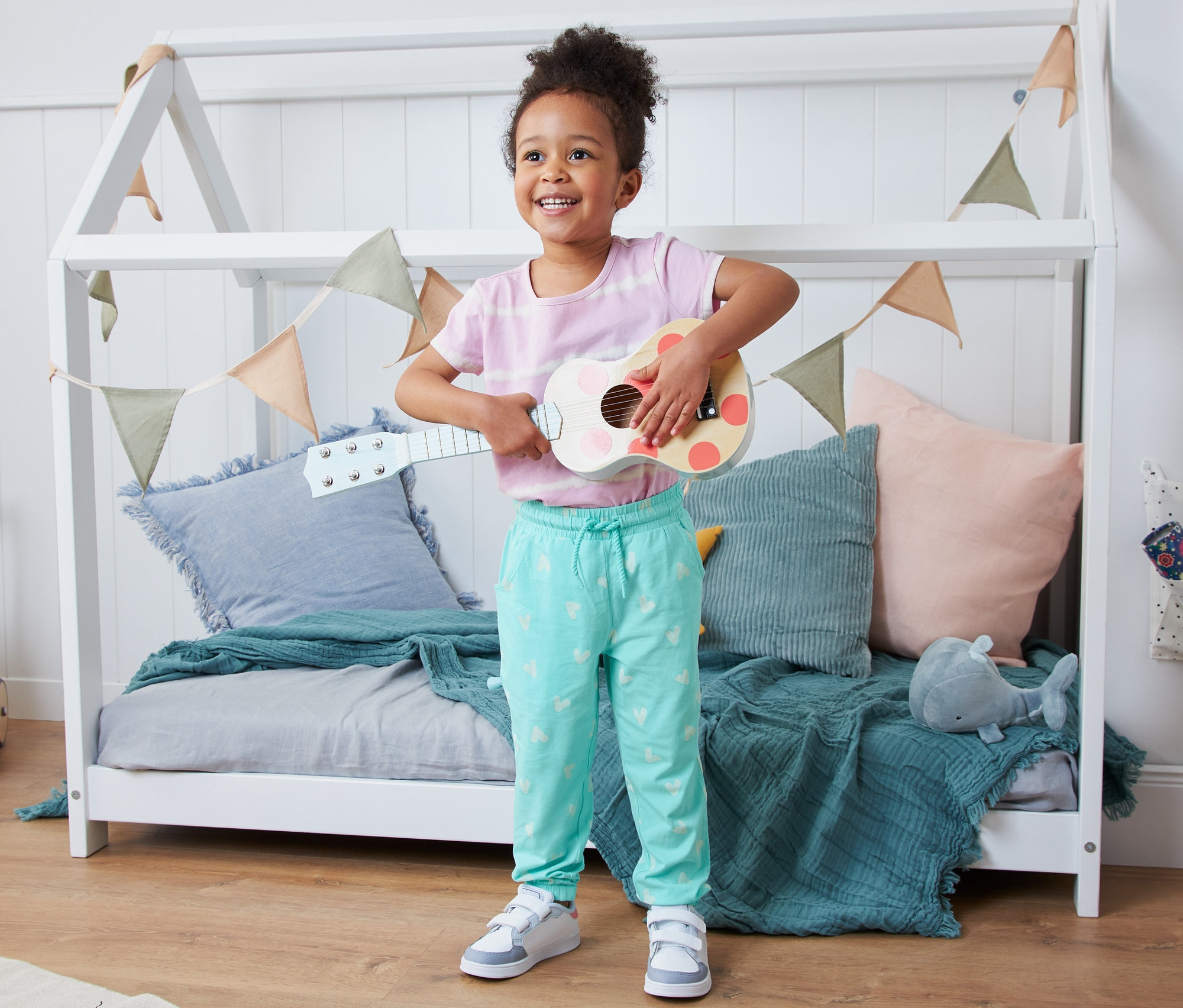 Lächelndes kleines Mädchen spielt in einem Kinderzimmer Ukulele. Sie trägt eine Schlupfhose aus Bio-Baumwolle.