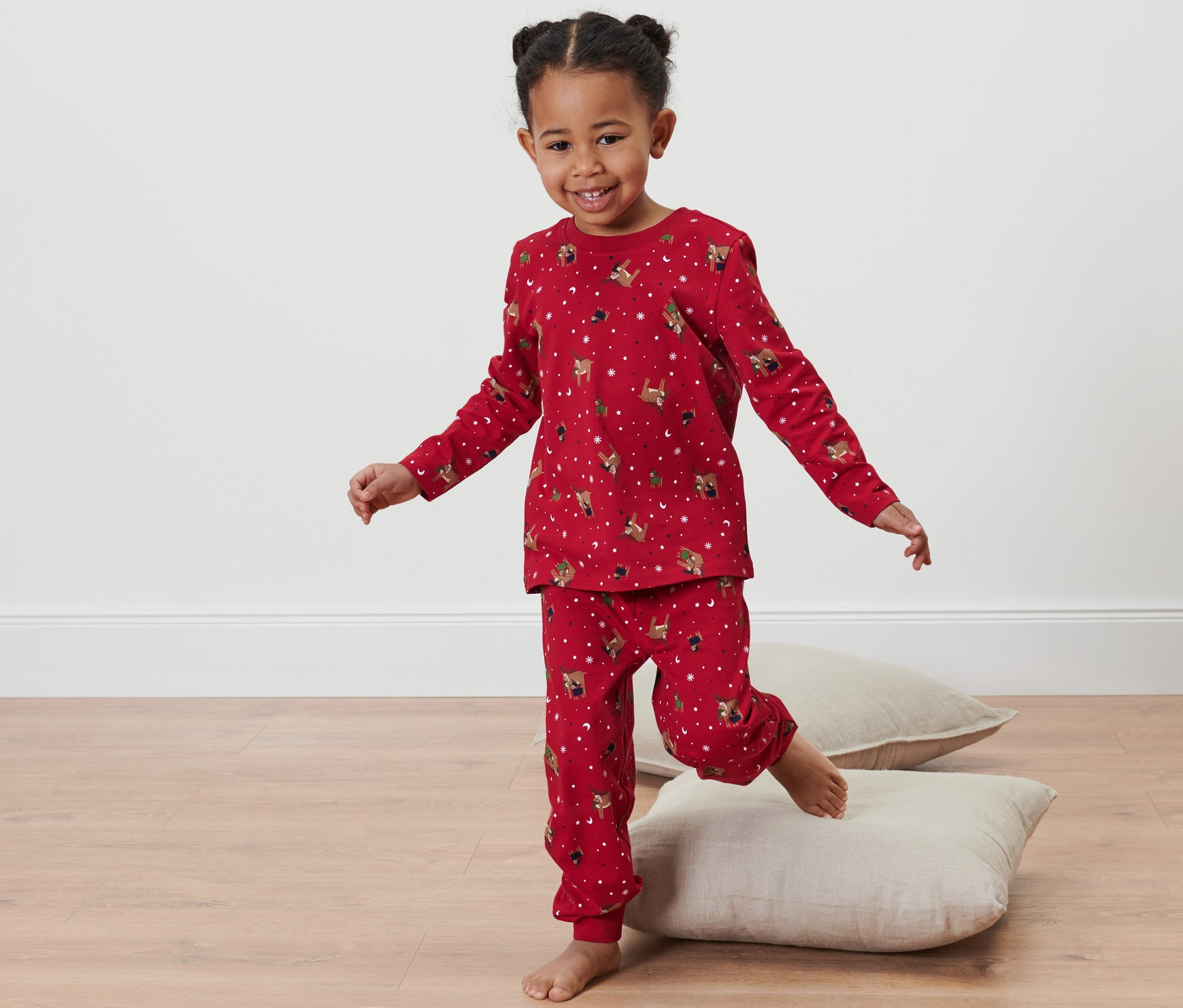 Ein Mädchen balanciert in einem roten Kleinkind-Jersey-Pyjama auf Kissen.