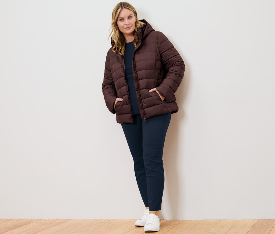 Eine Frau steht und trägt ein dunkelblaues Shirt mit 3/4-Arm und eine dunkelbraune Steppjacke mit Kapuze.