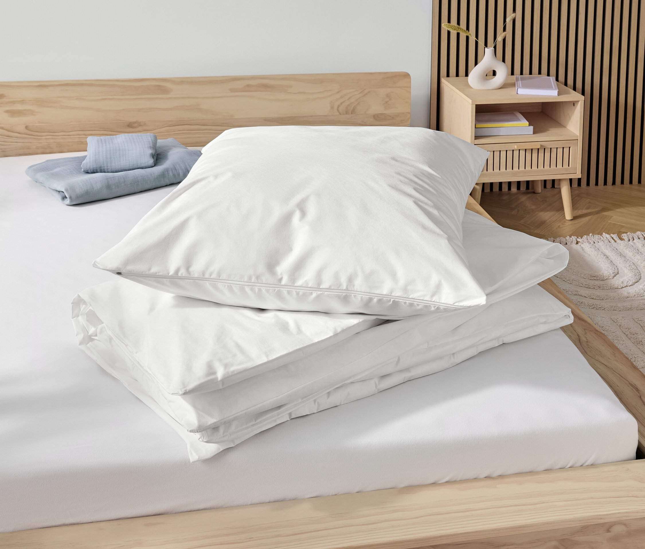Auf einem Holzbett liegen ein weißer Anti-Milben-Bettschutzbezug in Normalgröße und eine blaue Decke.
