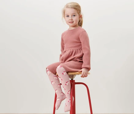 Mädchen sitzt auf rotem Hocker und trägt rosa Kleid und 5 Baumwoll-Strumpfhosen mit Einhorn.