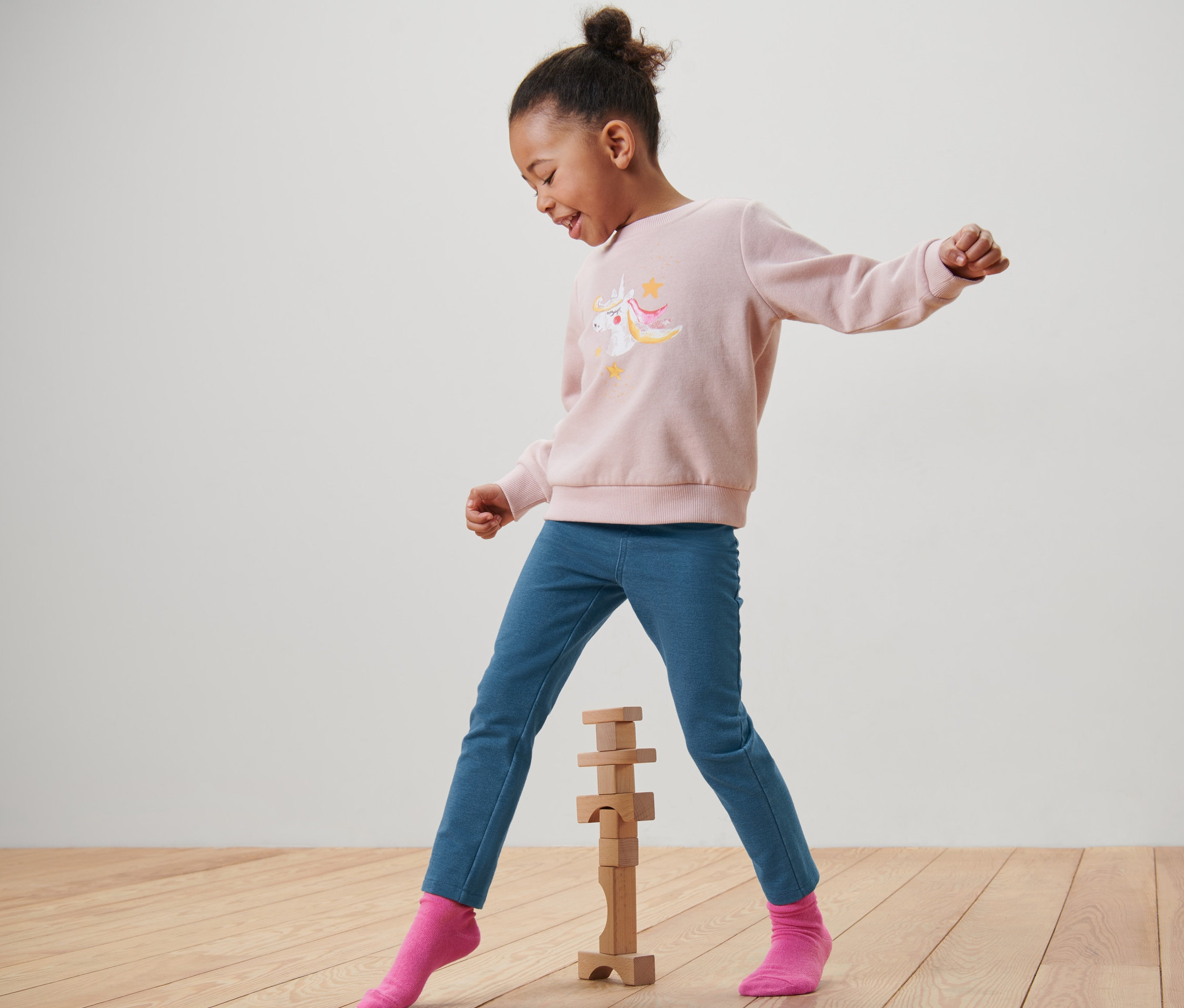 Ein Mädchen in einem rosa Einhorn-Sweatshirt und blauen »Mia« Kinder-Jeggings spielt mit Holzbausteinen.
