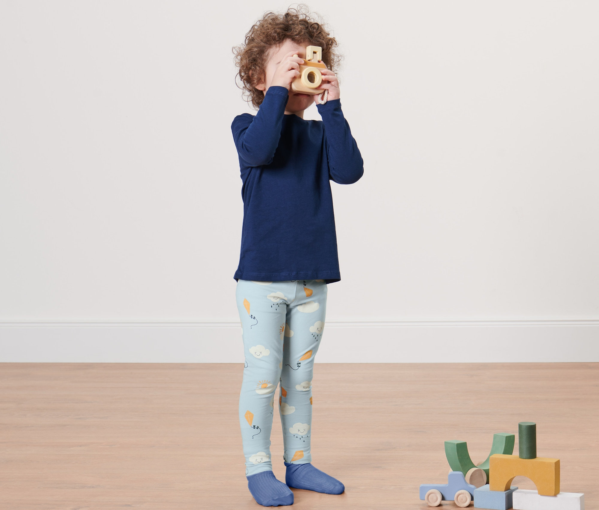 Ein Junge in blauem Oberteil und 2 Kleinkinder-Leggings spielt mit einer Holzkamera.