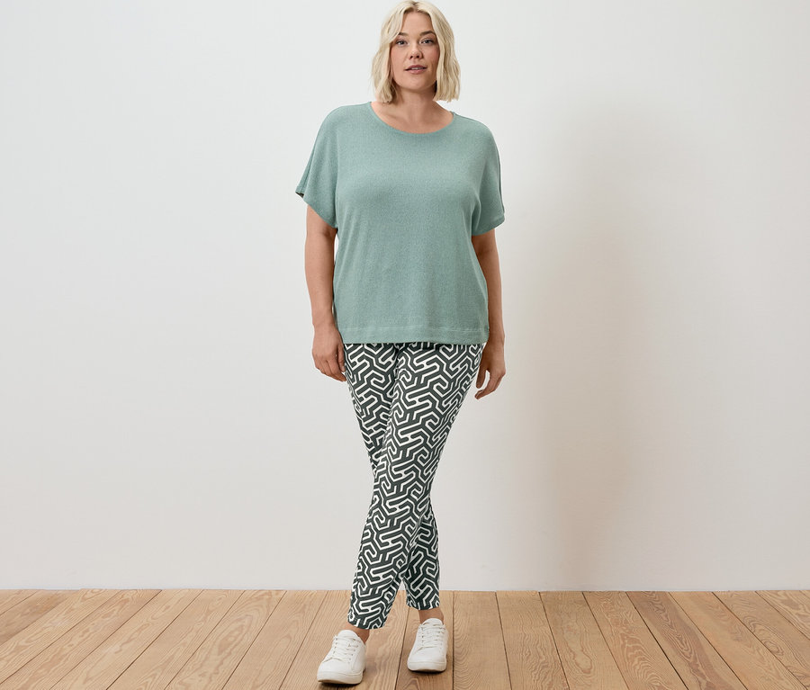 Frau steht und trägt ein grünes, strukturiertes Shirt und eine grüne, bedruckte Jerseyhose.