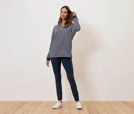 Eine Frau steht auf einem Holzboden und trägt ein gestreiftes Oberteil, dunkelblaue Leggings und weiße Turnschuhe.