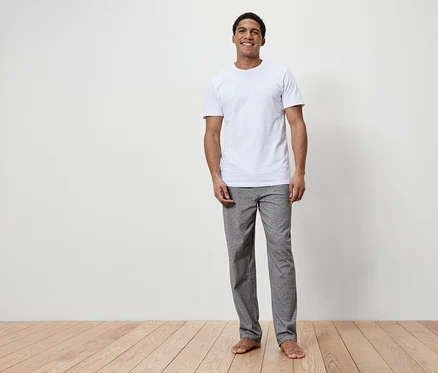 Ein lächelnder Mann steht barfuß auf einem Holzboden. Er trägt ein weißes T-Shirt und graue Hosen.