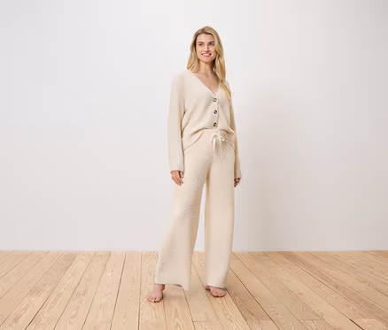 Blonde Frau steht in einem cremefarbenen Cardigan und Hose auf einem Holzboden.