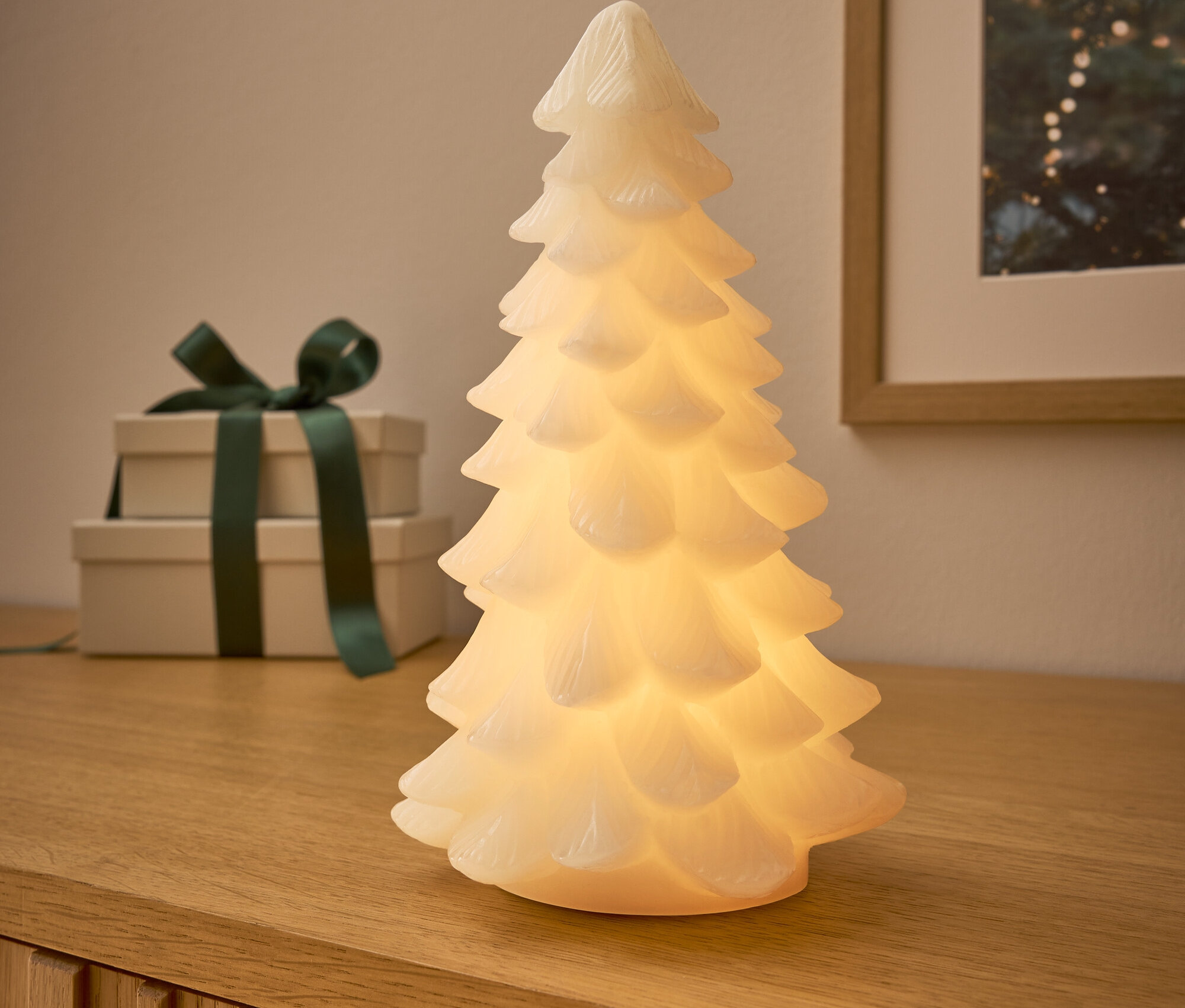 Auf einer Holzkommode stehen ein leuchtender Baum, dahinter zwei Geschenke mit dunkelgrünem Band und ein Bild mit Weihnachtsmotiv.