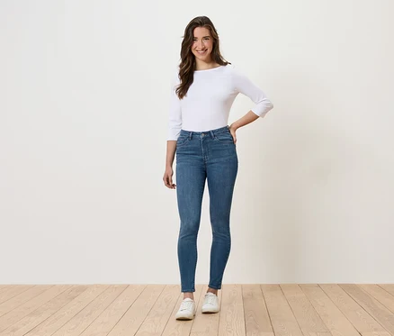 Eine Frau steht und posiert in blauen Jeans, einem weißen Oberteil und weißen Schuhen.