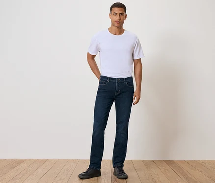 Ein Mann steht mit der Hand in der Tasche und trägt ein weißes T-Shirt, Jeans und schwarze Schuhe.