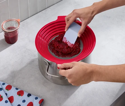 Eine Person passiert rote Früchte mit einem 2-in-1: Mehl-und-Passiersieb in einen Topf. Daneben steht ein Glas Marmelade und ein Geschirrtuch mit Erdbeermuster.