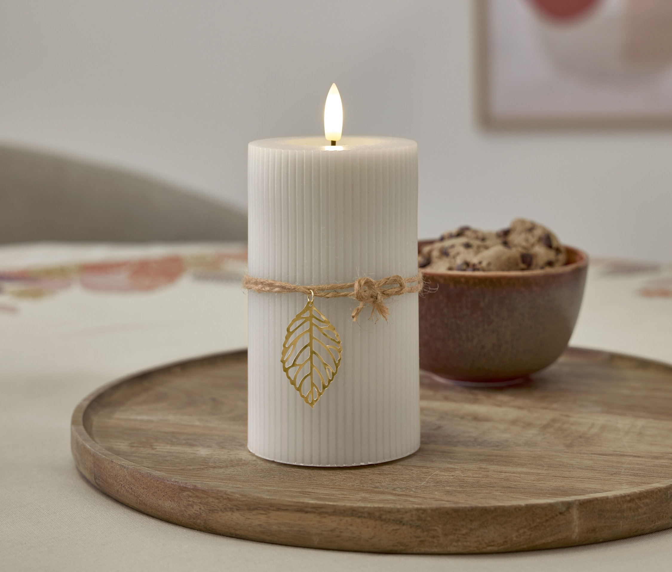 Auf einem Holztablett steht eine weiße Kerze mit Flamme, die mit einer Kordel mit Blattanhänger umwickelt ist, daneben eine Schüssel mit Keksen.