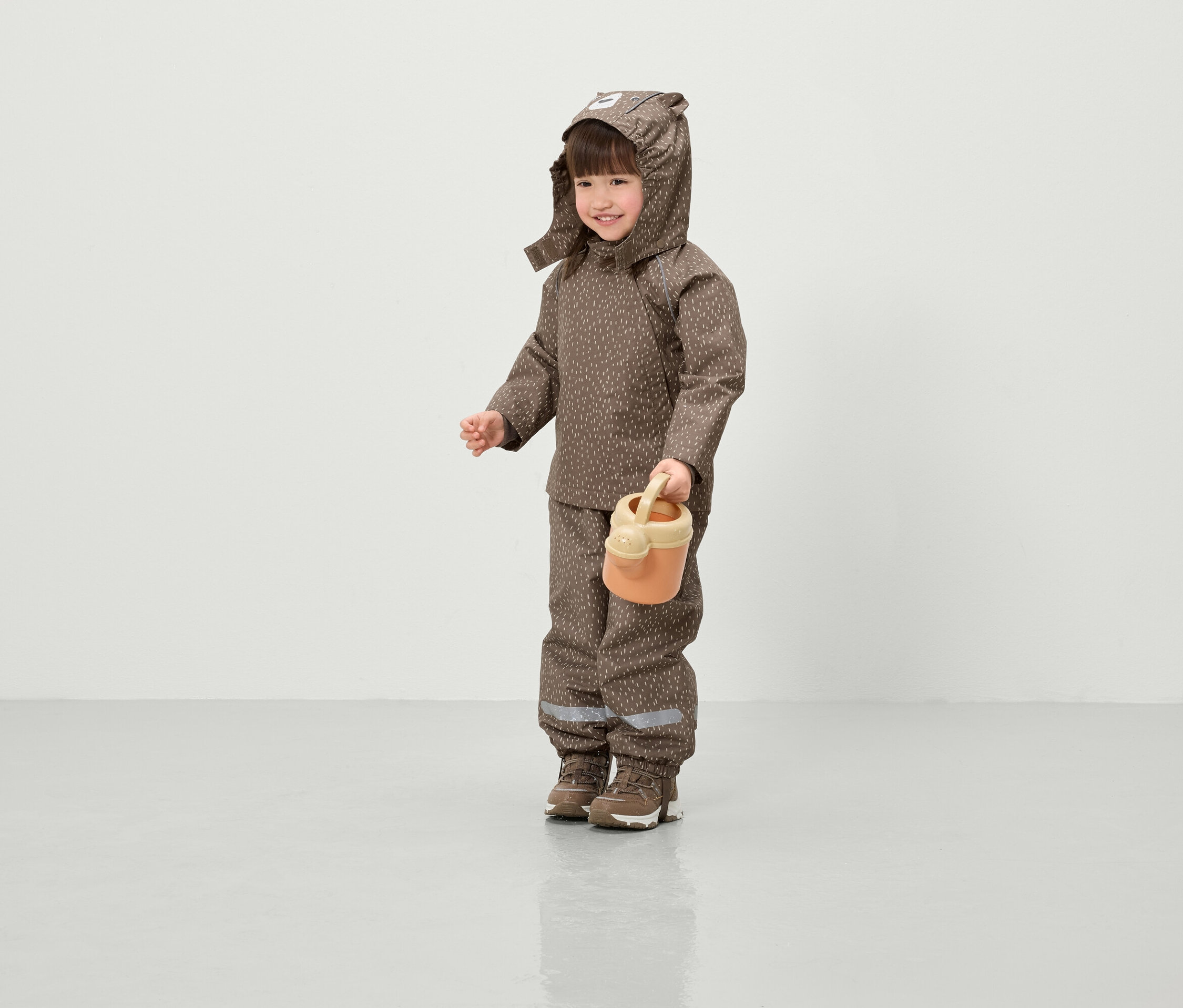 Ein lächelndes Kind in einer braunen, bedruckten Jacke mit Kapuze und Hose hält eine orangefarbene Gießkanne.