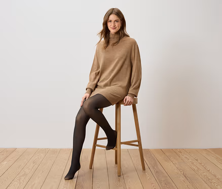 Frau sitzt auf einem Holzhocker, trägt ein braunes Rollkragenkleid und eine schwarze Strumpfhose.