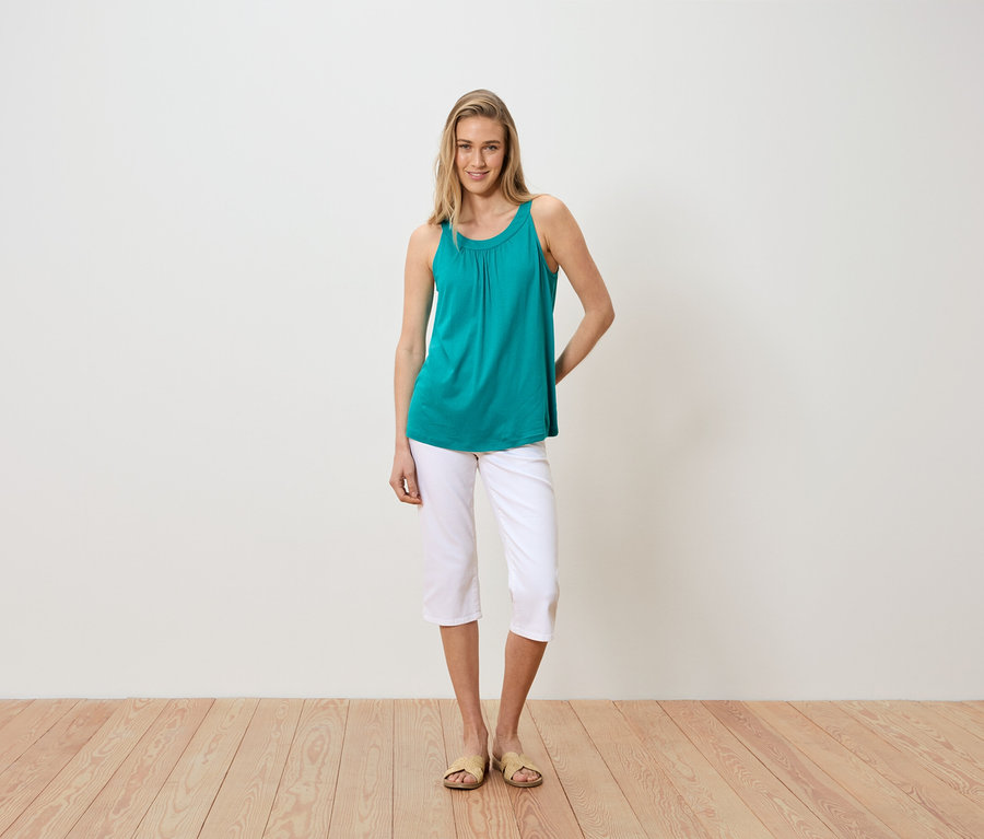 Eine Frau steht vor einer weißen Wand und trägt ein türkisfarbenes Tanktop, weiße Caprihosen und Sandalen.