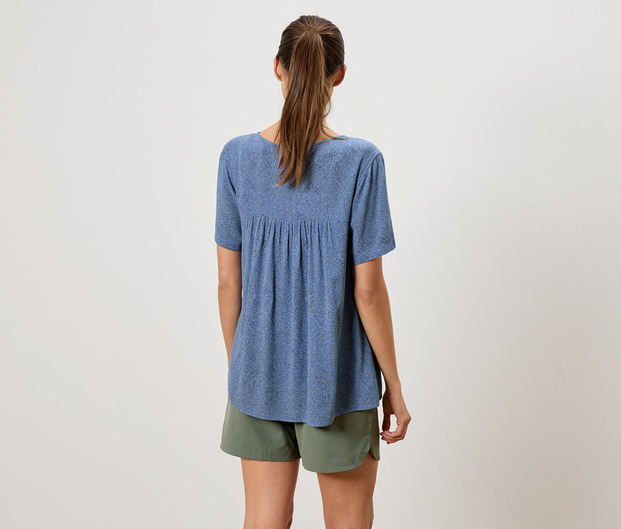 Rückansicht einer Frau mit Pferdeschwanz, die eine blaue, kurzärmlige Bluse und Shorts in Olivgrün trägt.
