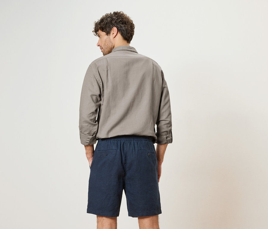 Ein Mann mit lockigem Haar steht mit dem Rücken zur Kamera, trägt ein graues Hemd und blaue Shorts.