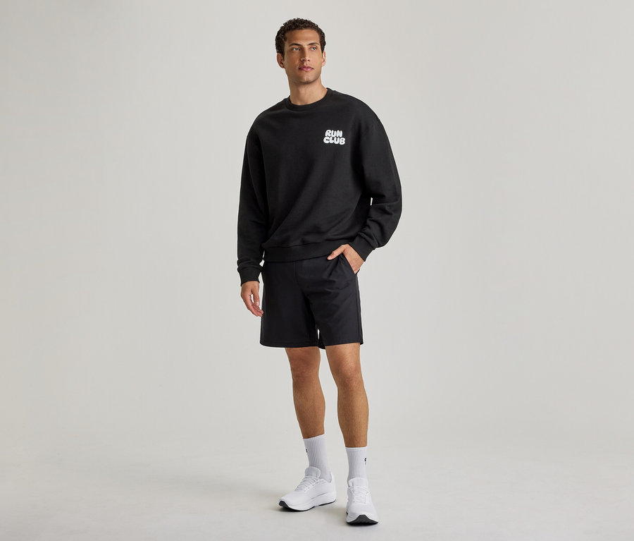 Ein Mann trägt ein schwarzes Sweatshirt mit der Aufschrift „RUN CLUB“, schwarze Shorts und weiße Turnschuhe. Seine linke Hand steckt in der Hosentasche.