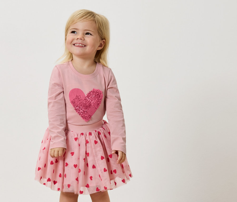 Ein kleines Mädchen mit blonden Haaren trägt ein rosa Langarmshirt mit einem großen, rosafarbenen Herz und einen rosa Tüllrock mit roten Herzen.