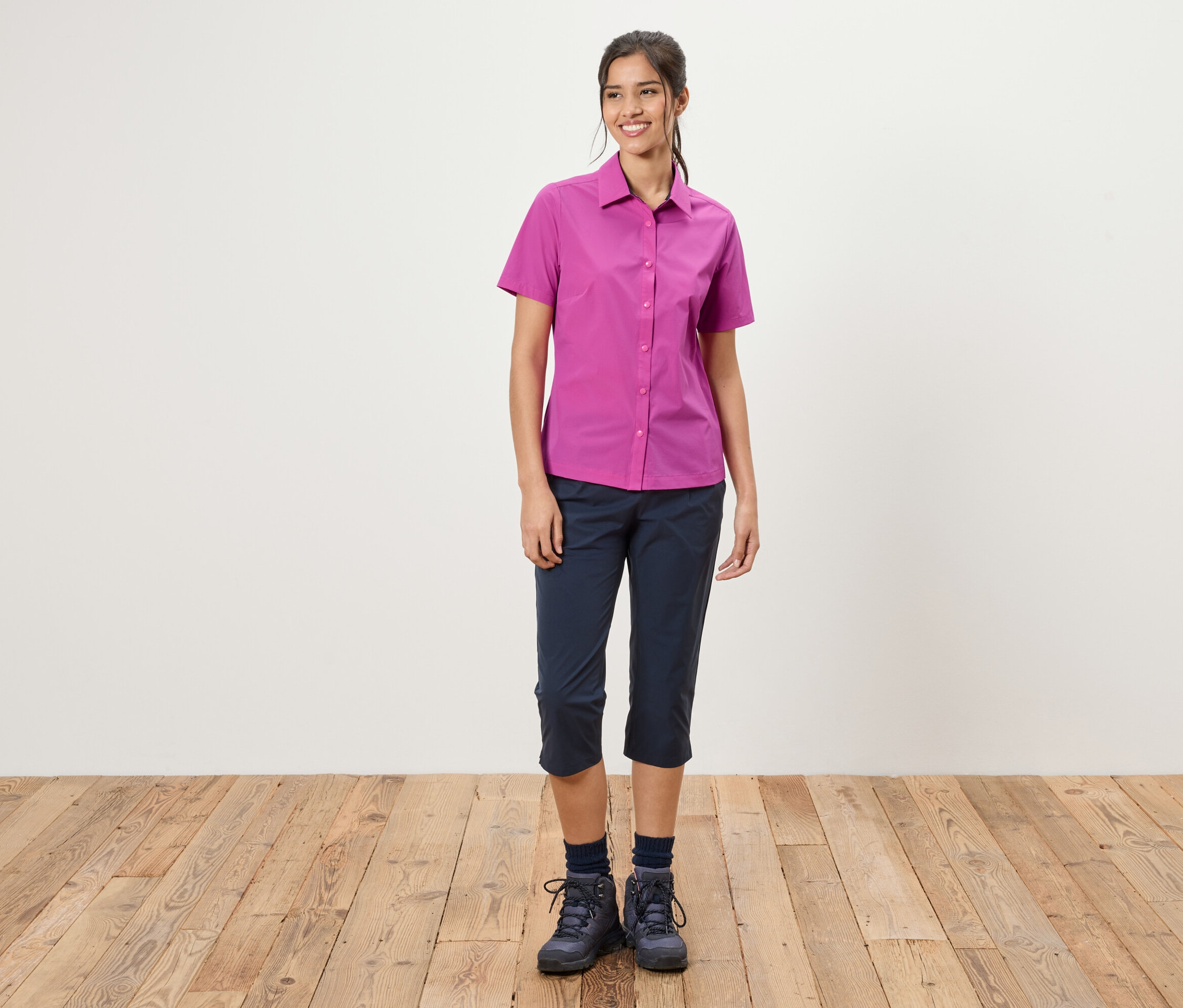 Eine Frau steht vor einer weißen Wand. Sie trägt ein pinkes Hemd, blaue Dreiviertelhosen und Wanderschuhe.