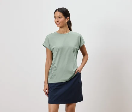 Frau mit hellgrünem T-Shirt mit Blumenmuster und blauem Rock, eine Hand in der Rocktasche.