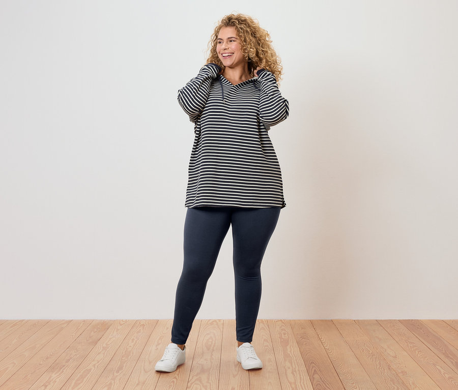 Frau mit lockigem Haar, trägt ein gestreiftes Oberteil, Leggings und weiße Turnschuhe.