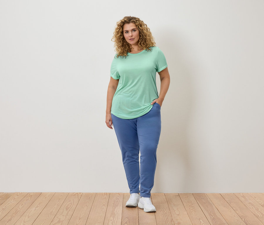 Frau mit lockigem Haar, trägt ein hellgrünes T-Shirt und blaue Jogginghose, steht auf einem Holzboden.