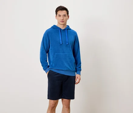 Ein Mann steht mit einer Hand in der Hosentasche vor einem neutralen Hintergrund. Er trägt einen blauen Kapuzenpullover und kurze, dunkelblaue Hosen.
