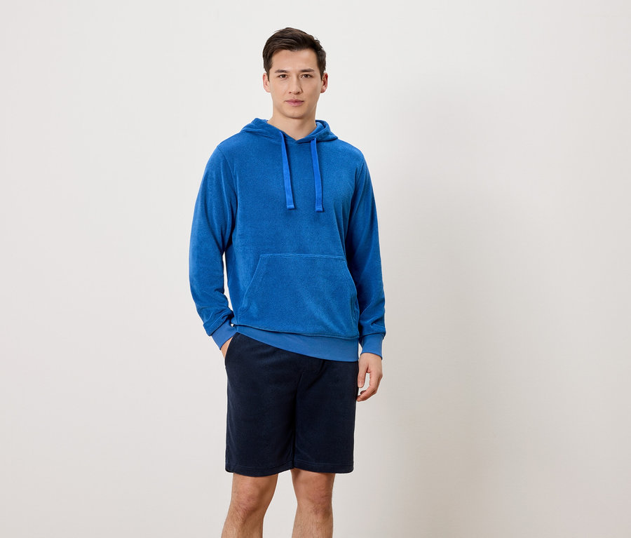 Ein Mann steht mit einer Hand in der Hosentasche vor einem neutralen Hintergrund. Er trägt einen blauen Kapuzenpullover und kurze, dunkelblaue Hosen.