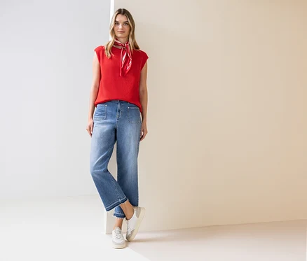 Eine Frau steht und trägt ein rotes Top, ein Halstuch, Jeans und Turnschuhe.