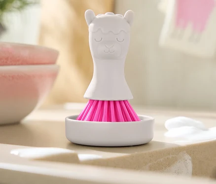 Eine weiße Bürste mit rosa Borsten in Form eines Lamas steht in einer weißen Schale auf einer Spüle mit Seifenresten.
