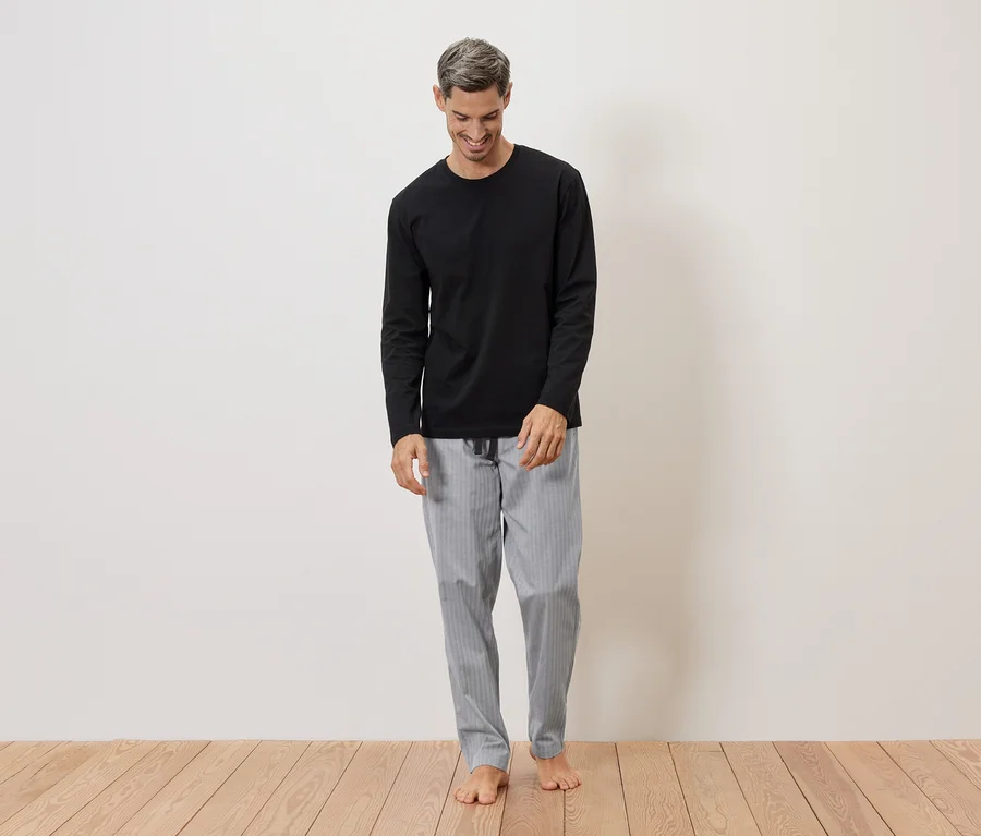 Mann mit grauen Haaren trägt ein schwarzes Langarmshirt und eine grau gestreifte Pyjamahose, barfuß vor einer weißen Wand.