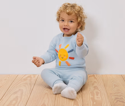 Ein lächelndes Baby mit blonden Locken sitzt auf einem Holzboden und trägt einen hellblauen Pullover mit einer aufgestickten Sonne und eine passende Hose.