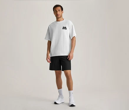 Ein Mann steht nach vorne blickend und trägt ein weißes Sportshirt mit dem schwarzen Schriftzug „RUN CLUB“ auf der Brust, schwarze Shorts, weiße Socken und weiße Sportschuhe.