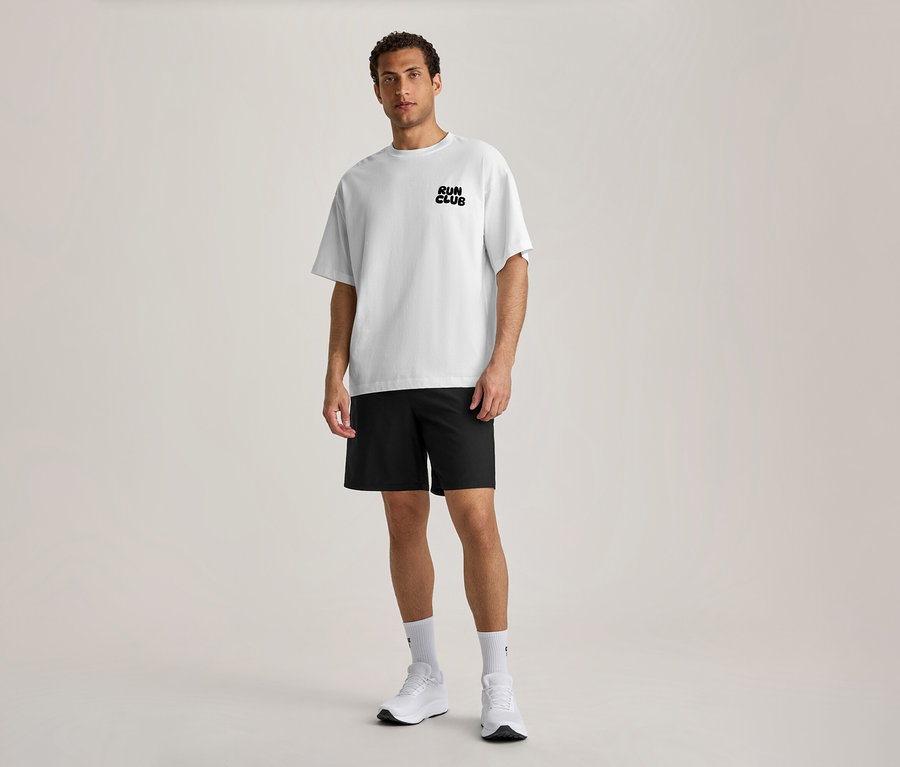 Ein Mann steht nach vorne blickend und trägt ein weißes Sportshirt mit dem schwarzen Schriftzug „RUN CLUB“ auf der Brust, schwarze Shorts, weiße Socken und weiße Sportschuhe.