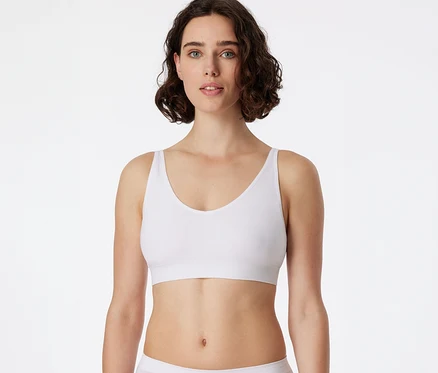 Frau mit kurzen, dunklen Locken trägt einen weißen SCHIESSER Soft BH mit Pads und blickt in die Kamera.