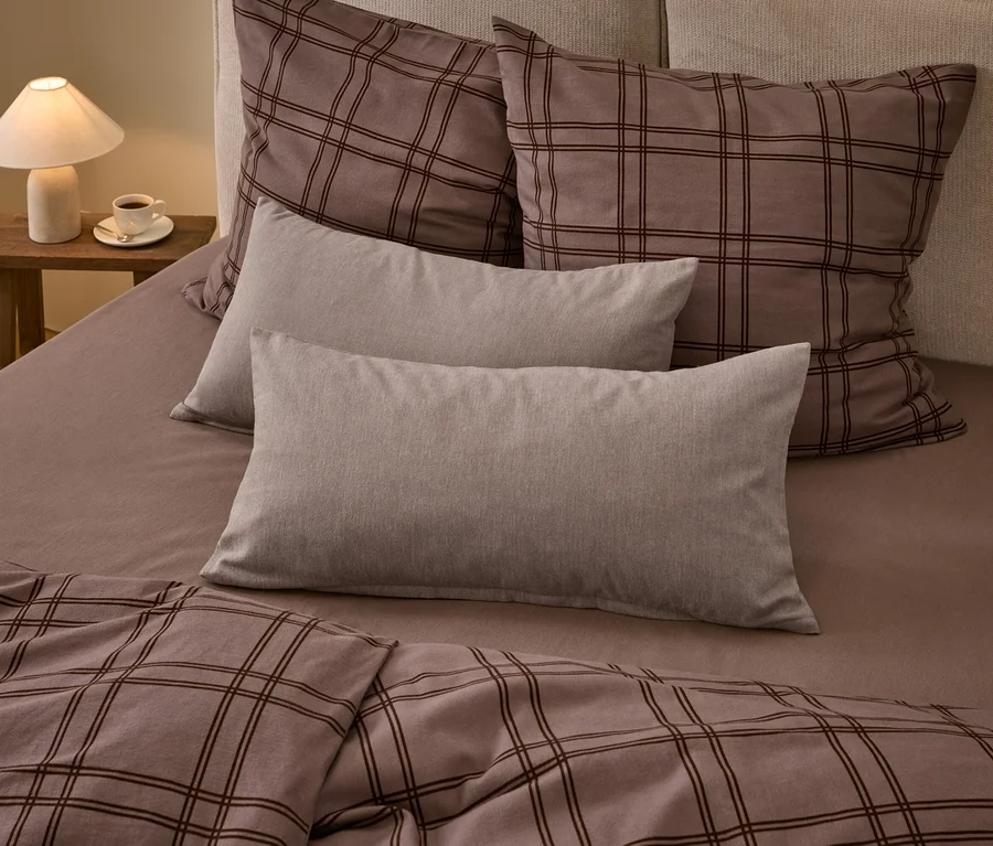 Ein Bett mit brauner karierter Bettdecke und zwei passenden großen Kissen, zwei einfarbigen hellgrauen rechteckigen Kissen und einem braunen Spannbettlaken. Eine weiße Lampe und eine Tasse Kaffee stehen auf einem Holznachttisch.