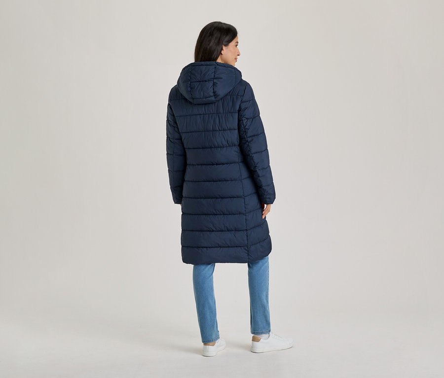 Frau steht mit dem Rücken zum Betrachter, trägt einen langen, marineblauen Steppmantel mit Kapuze, blaue Jeans und weiße Turnschuhe.