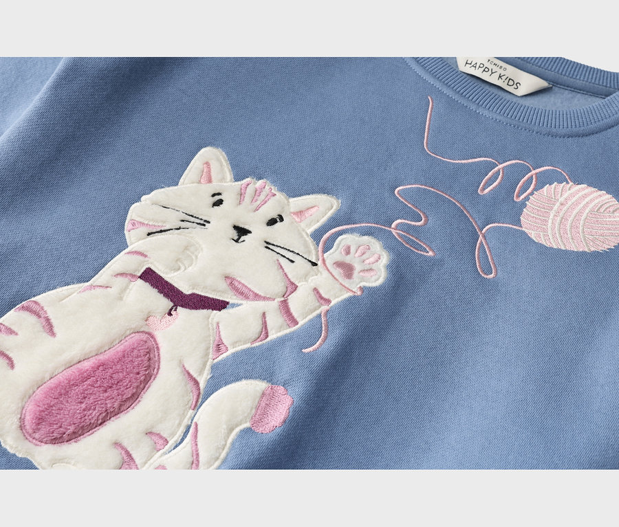Nahaufnahme eines blauen Sweatshirts mit einer applizierten weißen Katze mit rosa Streifen und flauschigem rosa Bauch, die nach einem gestickten rosa Garnfaden und Wollknäuel greift.
